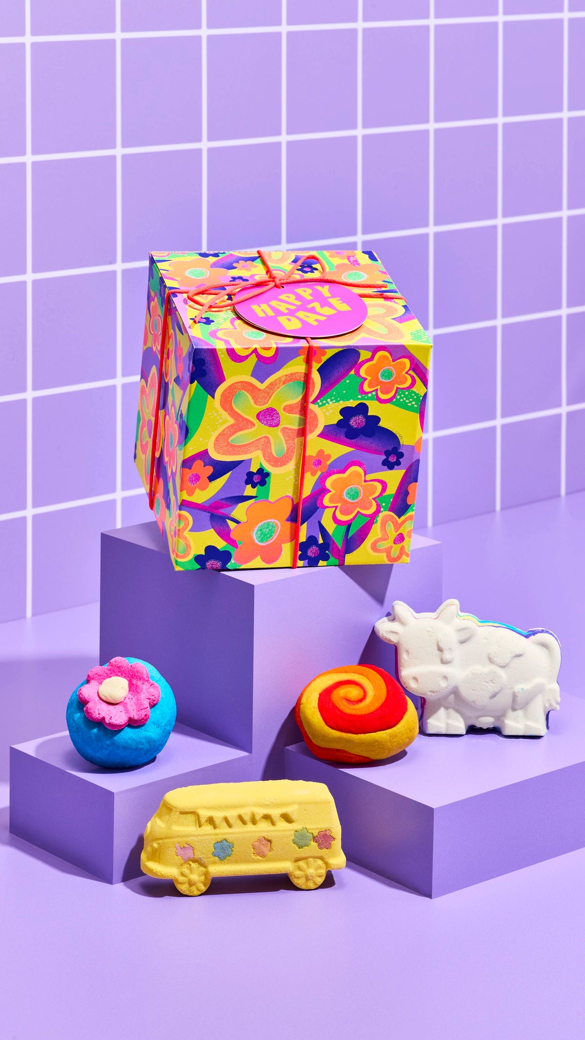 The image shows the colourful neon flower-designed Happy Daze gift box, sitting on a cubed platform with the four included bath products sitting on lower platforms on a pastel-purple tiled background.