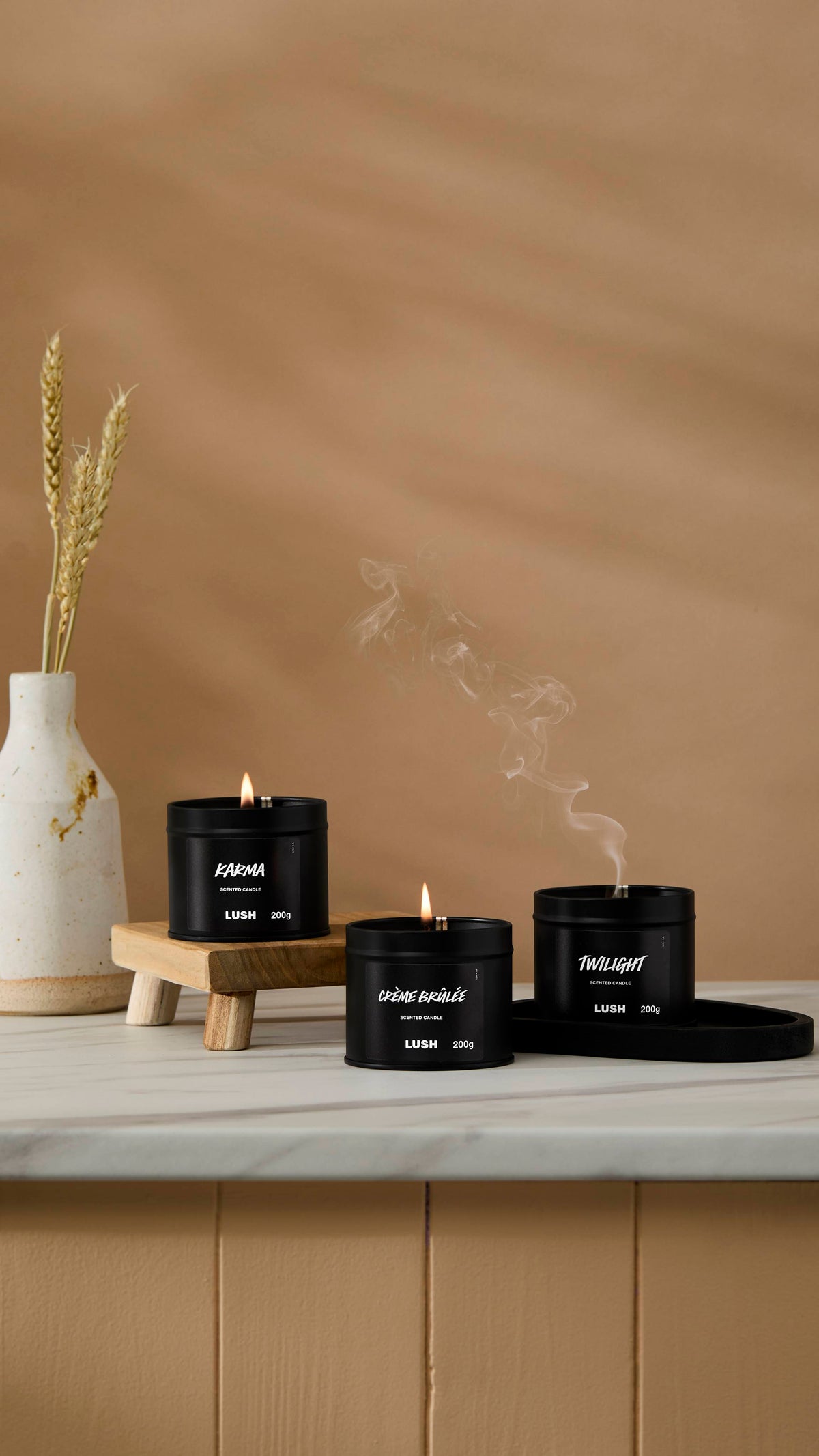 The image shows a warm beige background with a ceramic vase of wheat. Three back tin candles are sitting with their wicks lit on a marble-topped counter.
